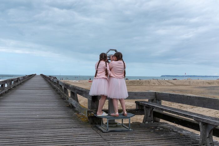 Two girls in pink dresses on a wooden pier capturing moments, showcasing street photography at a beach setting.