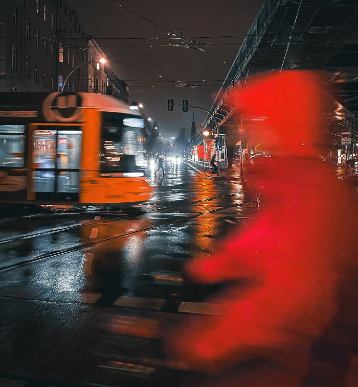 Blurred figure in red coat crossing wet street at night with a moving tram, captured in street photography awards image.