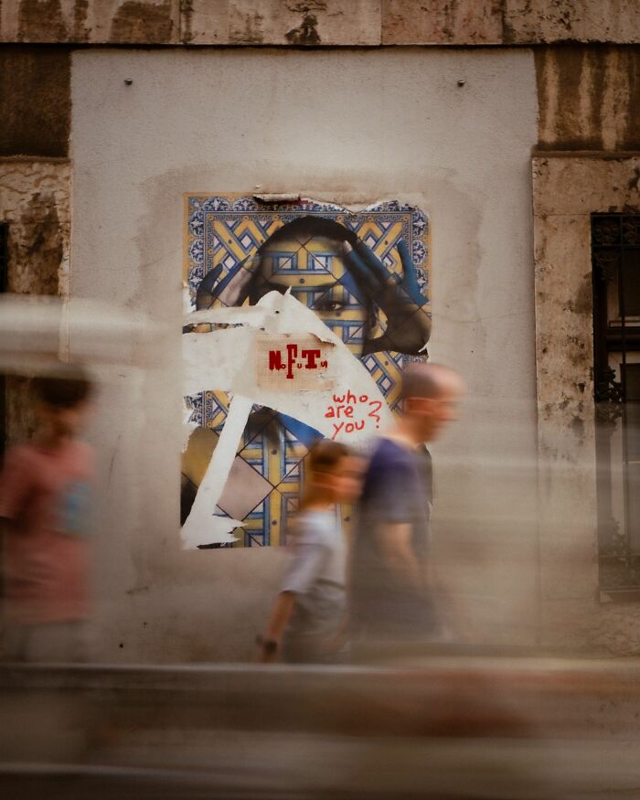 Blurred pedestrians walk past street art featuring a partially torn poster with the words who are you in urban street photography.