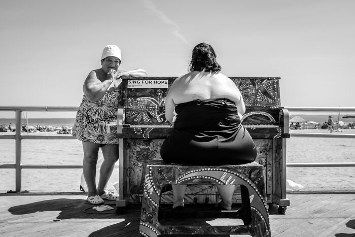 Black and white street photography of two women by a decorated piano on a boardwalk, capturing raw moments in pure street photography.