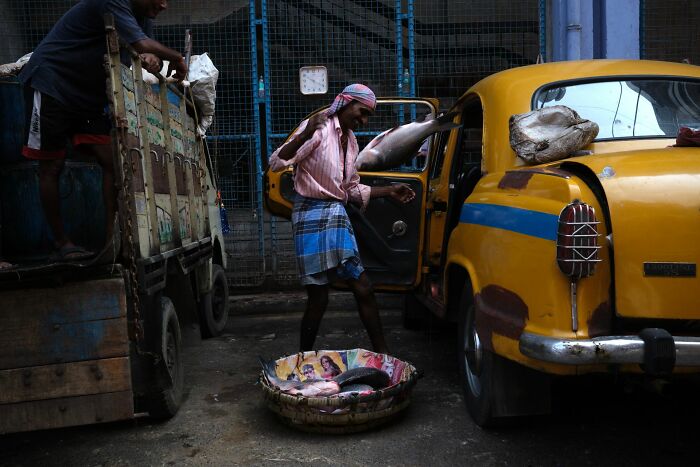 Man unloading fish near a yellow car in a dimly lit street, captured in Pure Street Photography Awards 2025.