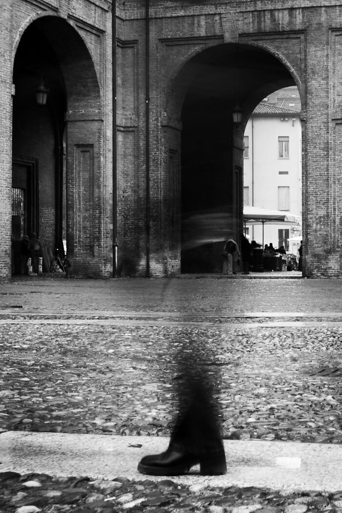 Black and white street photography showing blurred pedestrian walking past historic brick arches in urban setting.