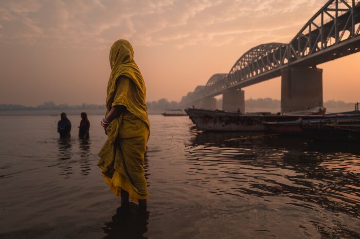 Woman in yellow sari standing in water near boats at sunset, captured in pure street photography awards scene.