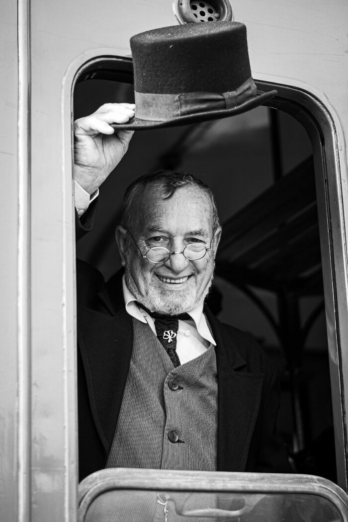 Older man smiling and tipping his hat inside a vintage train carriage, classic style street photography portrait.
