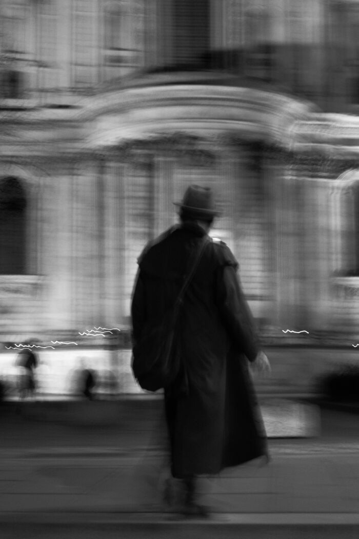 Blurry black and white street photography of a person in a long coat and hat walking past historic architecture.