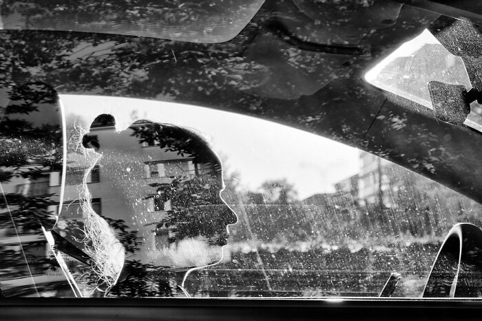 Black and white pure street photography capturing a woman’s silhouette through a car window with urban reflections and light.