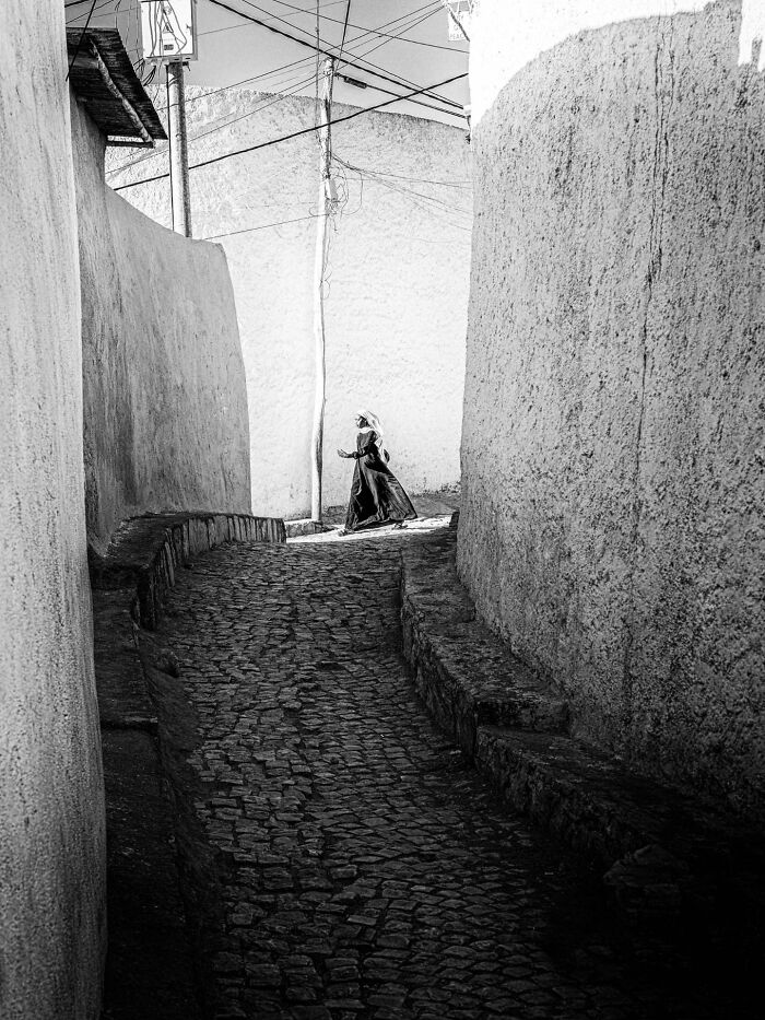 Woman in flowing dress walking on cobblestone path between white walls in pure street photography awards image.