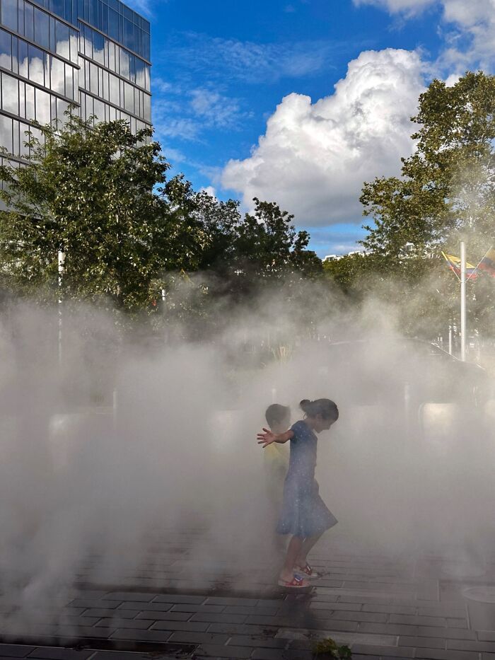 Child playing in mist on city street with trees and building in background, pure street photography awards theme.