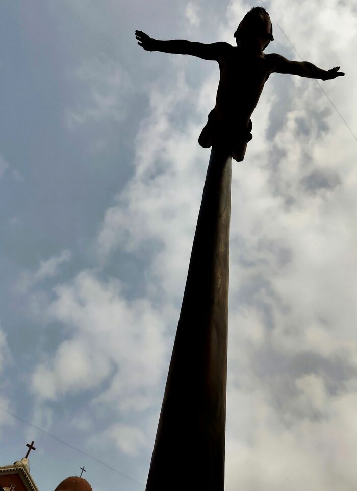 Silhouette of a child with arms outstretched atop a tall pole against a cloudy sky, street photography award entry.