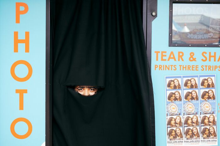Person peeking through a black curtain inside a photo booth, capturing a candid moment in street photography.