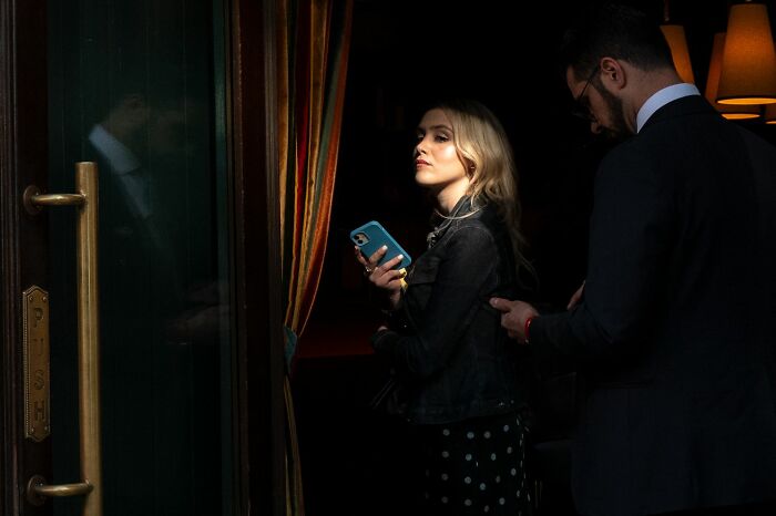 Young woman holding a phone and a man with glasses in a dark urban setting, capturing pure street photography vibes.