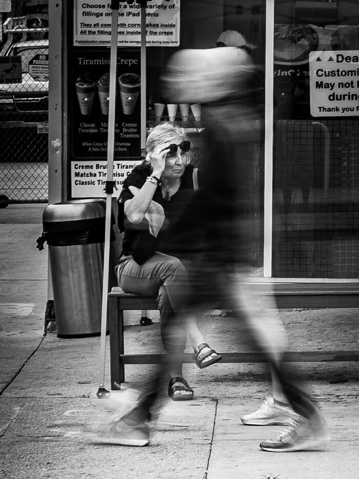 Black and white street photography of a seated woman adjusting sunglasses with blurred pedestrian walking by.