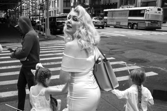 A woman in a white dress holding two children’s hands while crossing the street in urban street photography.