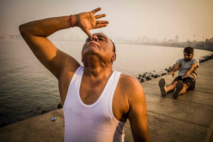 Man in white tank top stretching by waterfront with another person exercising on pathway in pure street photography awards scene