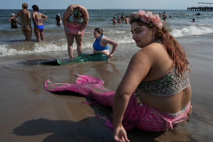 A group of people at the beach with two women dressed as mermaids, capturing a candid moment for street photography awards.