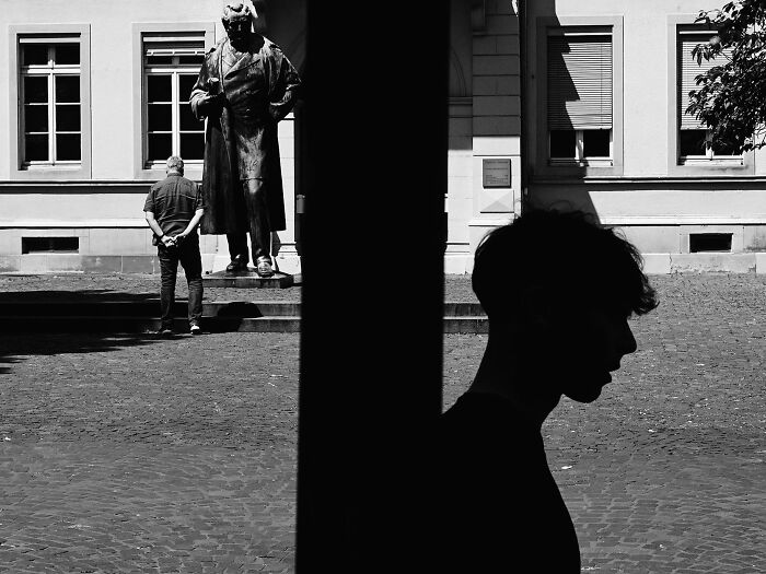 Black and white street photography showing a silhouette of a person opposite a man near a statue, pure street photography awards.