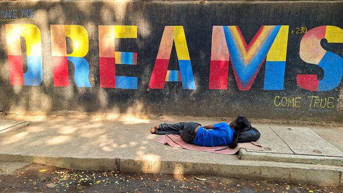Person lying on the sidewalk in front of a colorful street mural with the word dreams in pure street photography awards style.