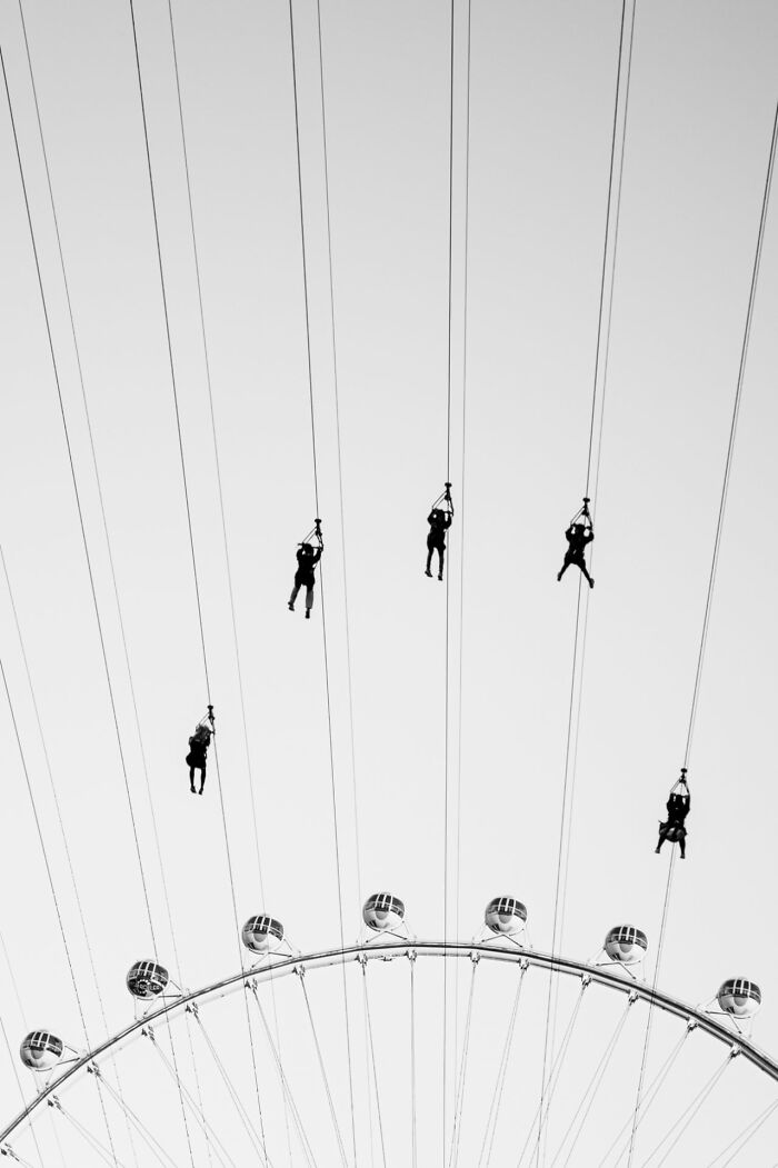 Silhouettes of people ziplining above a Ferris wheel captured in black and white street photography awards image.