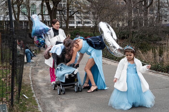 Street scene in a park showing a woman adjusting a stroller and a child in a blue dress holding balloons, pure street photography award theme.