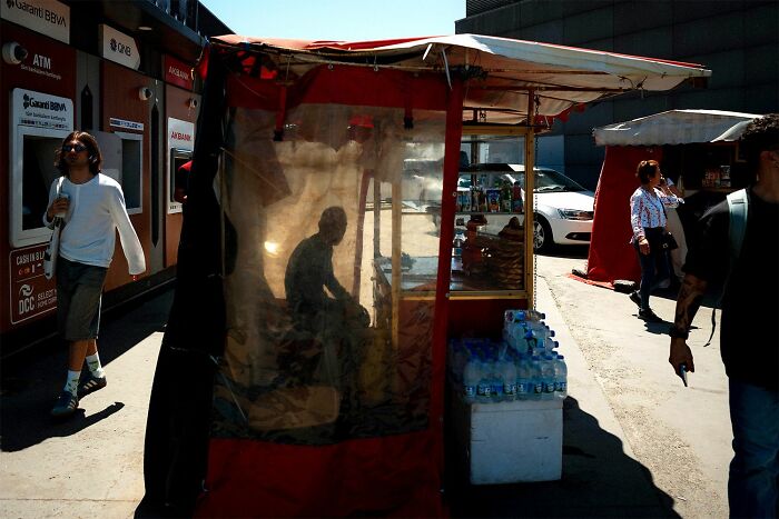 Shadowed figure sitting inside a street vendor stall with pedestrians walking by in bright daylight in street photography style