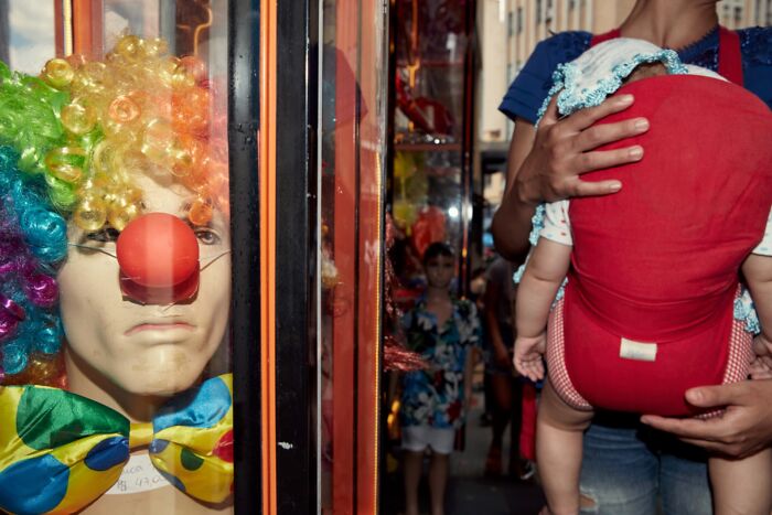 Colorful clown mannequin behind glass beside a person carrying a baby in a red carrier in street photography scene