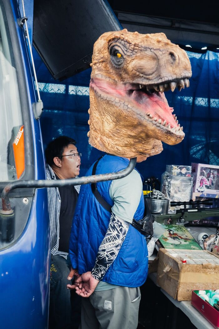 Man wearing dinosaur head mask and blue vest standing next to surprised man in a street market scene, street photography.