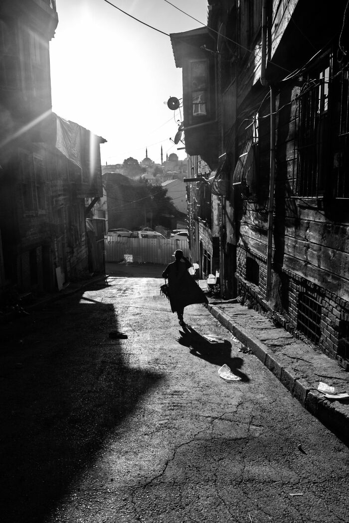 Black and white street photography showing a lone figure walking down a narrow sunlit urban street at sunset.