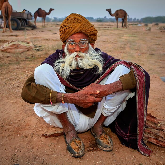 Elderly man in traditional attire sitting on dusty ground with camels in the background, captured in street photography awards.
