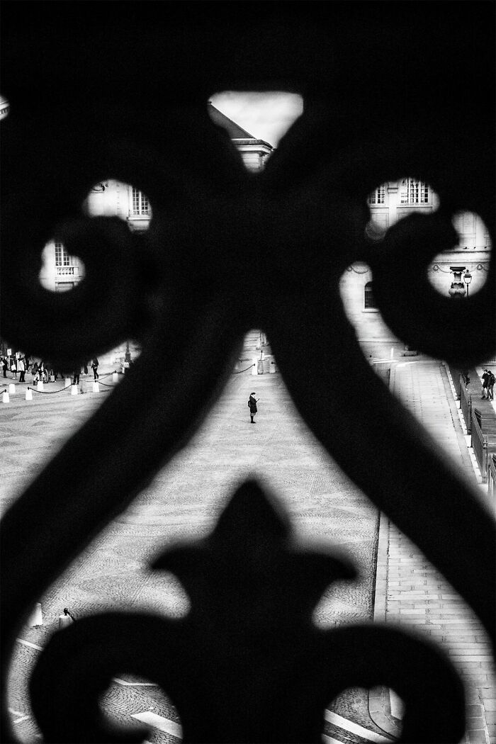Black and white street photography capturing a lone person through ornate ironwork in urban setting for Pure Street Photography Awards.