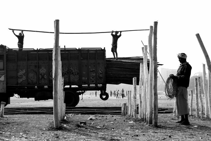 Black and white street photography showing workers loading poles onto a truck in an open urban area with scattered wooden posts.