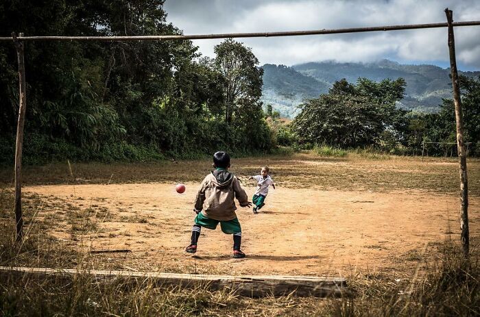 Two children playing soccer on a rustic dirt field surrounded by trees and mountains, capturing childhood moments worldwide.