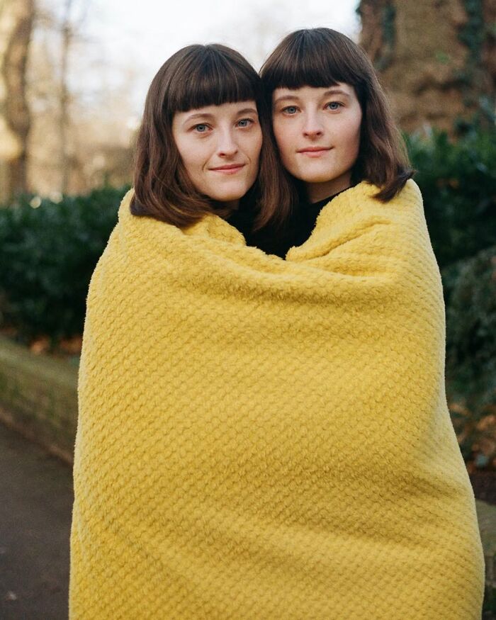 Twin sisters wrapped in a yellow blanket in an outdoor setting, captured by a London photographer in a stunning portrait.
