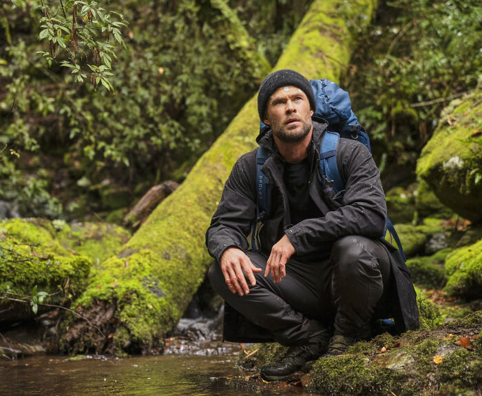 Man in outdoor gear sitting by a forest stream, exploring nature and reflecting on unknown things about celebrities.