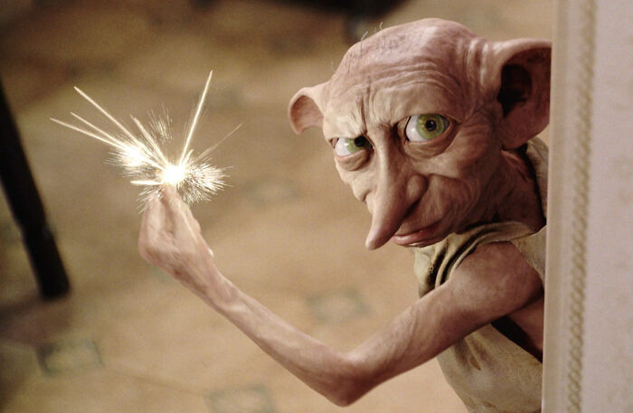 Dobby the house-elf from Harry Potter holding a glowing magical light at the tip of his finger, indoors.
