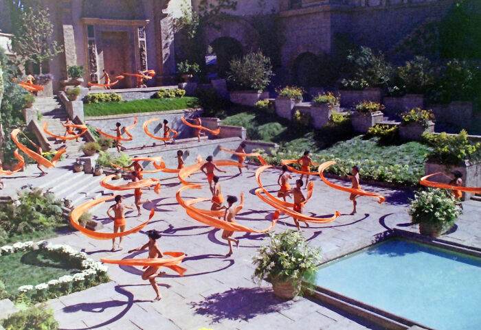 Dancers with flowing orange ribbons performing outdoors in a magical imaginary world with lush gardens and stone architecture.