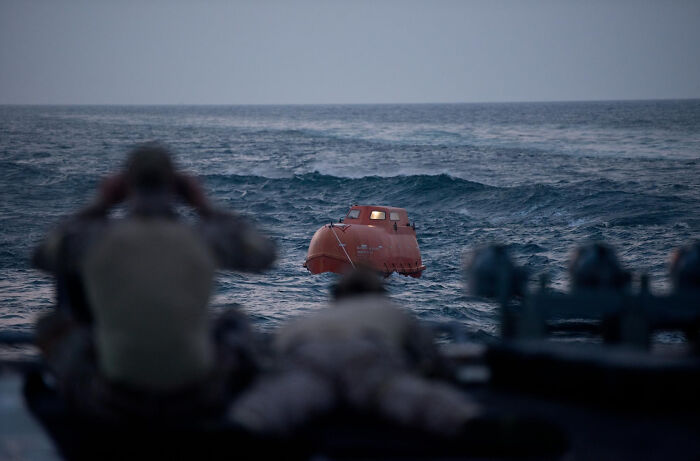 Orange lifeboat floating in rough ocean waves, with blurred figures watching from a nearby vessel, depicting true story events.