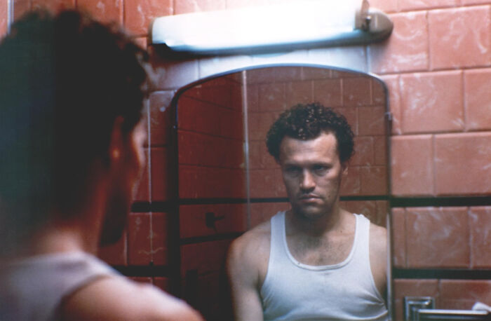 Man in a white tank top looking at his reflection in a bathroom mirror from a movie based on true stories that feel impossible.