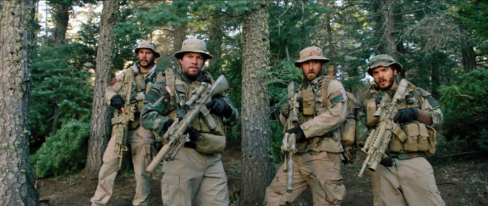 Four soldiers in tactical gear holding rifles, standing alert in a dense forest setting in a movie scene based on true stories.