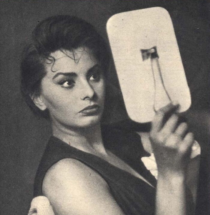 Sophia Loren looking into a handheld mirror, showcasing her Italian beauty and timeless Hollywood elegance in a black and white photo.