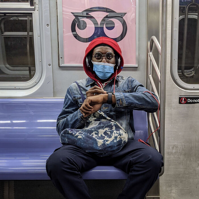 Person wearing glasses and a mask, sitting on a subway bench with headphones, captured in a NYC street portrait.