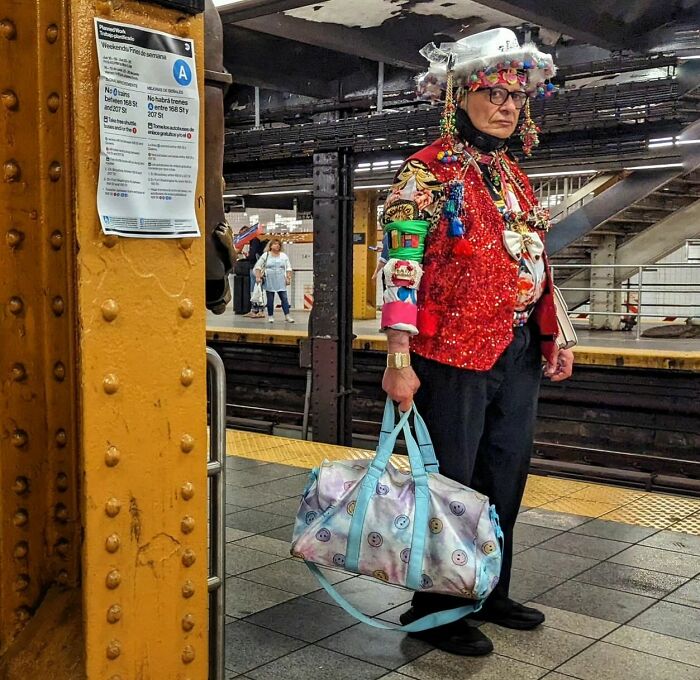 Colorfully dressed person with decorated hat and large bag standing on NYC subway platform in street portrait style.