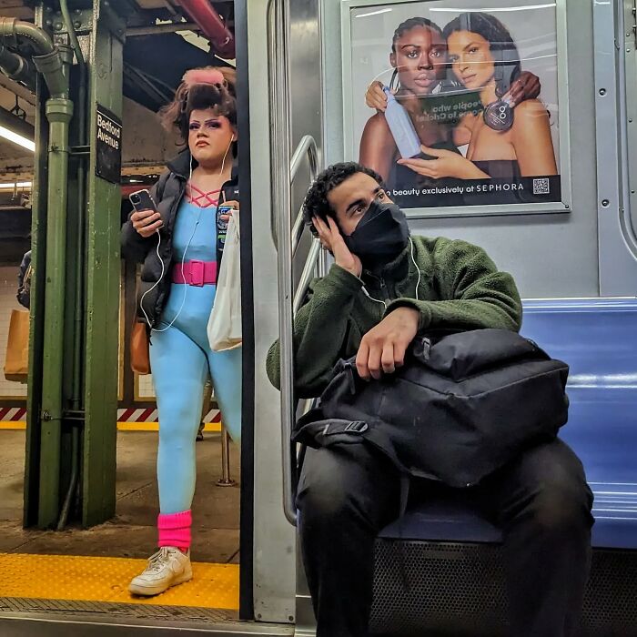 Woman in bright outfit waiting on subway platform as man wearing mask sits inside NYC subway car street portrait.