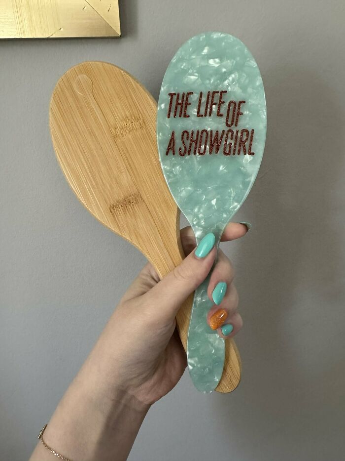 Hand holding a teal hairbrush with The Life of a Showgirl text alongside a wooden hairbrush, Taylor Swift merchandise.