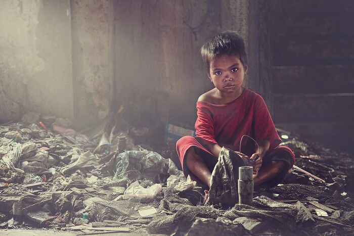 Childhood around the world depicted by a young child in a red shirt sitting among debris in a dimly lit space.