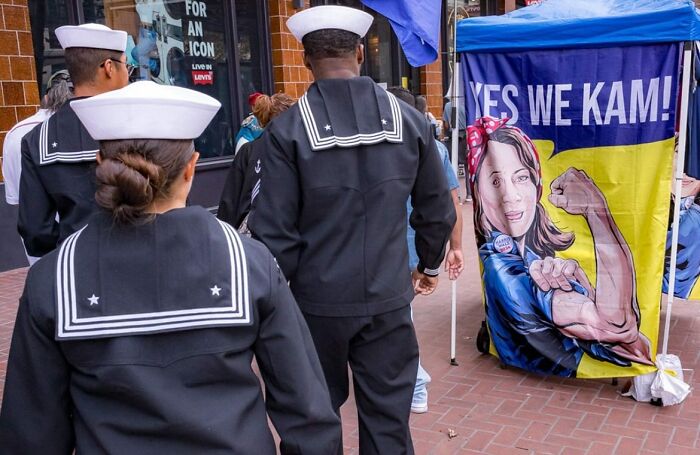 Street photographer captures humor and chaos as sailors walk past colorful political mural on busy city street.