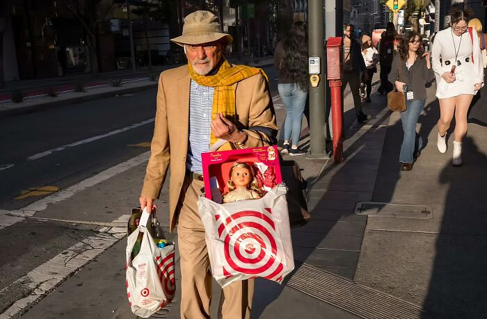 Street photographer captures humor and chaos in everyday life with a man carrying Target bags and a doll on a busy sidewalk.