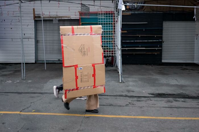 Person walking on street covered by large cardboard box, capturing humor and chaos in everyday life street photography.