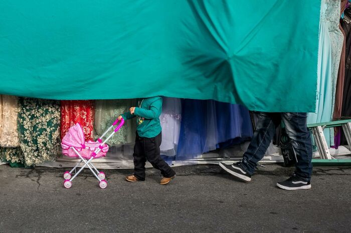 Child pushing a toy stroller beneath hanging fabric, capturing humor and chaos in everyday street photography.