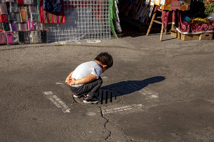 Young boy squatting over a street drain, captured by street photographer highlighting humor and chaos in everyday life.