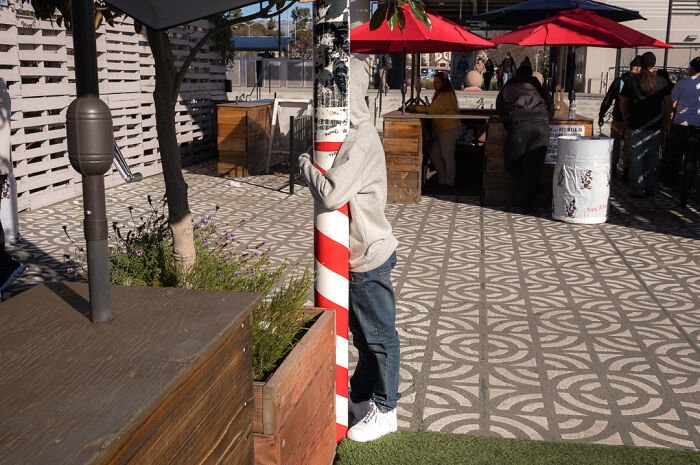 Street photographer captures humor and chaos as person hides behind a red and white striped pole in a busy outdoor area.